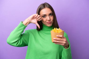 Mor arka planda kızarmış patates kızartması tutan genç beyaz kadın, olumsuz bir ifadeyle baş parmağını aşağı gösteriyor.