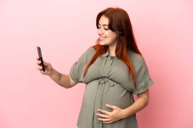 Genç kızıl saçlı beyaz kadın pembe arka planda izole edilmiş hamile ve cep telefonu kullanıyor.