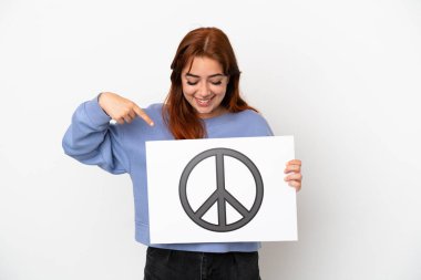 Genç kızıl saçlı kadın beyaz arka planda izole edilmiş. Elinde barış sembolü olan bir pankart var ve onu işaret ediyor.