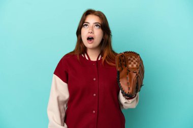 Beyzbol oynayan genç kızıl saçlı kadın mavi arka planda izole edilmiş yukarı bakıyor ve şaşırmış bir ifadeyle