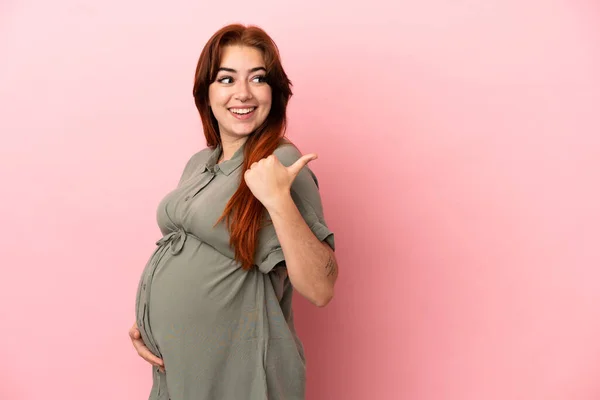 妊娠中のピンクの背景に隔離され 横を指している若い赤毛の白人女性 — ストック写真