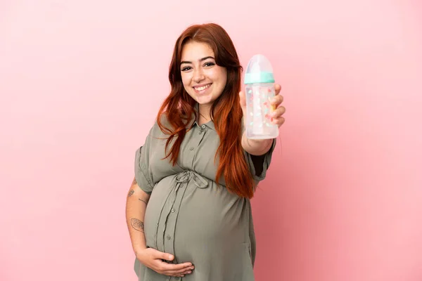 若い赤頭の白人女性はピンクの背景に孤立妊娠中や授乳瓶を保持 — ストック写真