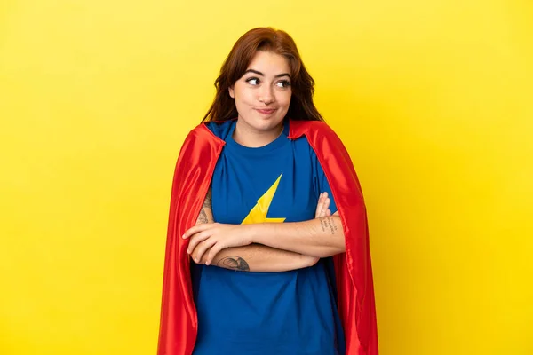 Super Hero redhead woman isolated on yellow background making doubts gesture while lifting the shoulders