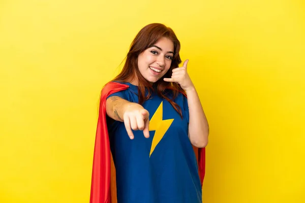 Super Hero redhead woman isolated on yellow background making phone gesture and pointing front