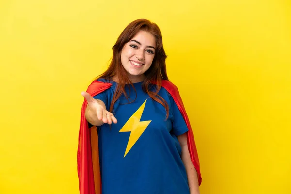 Super Hero redhead woman isolated on yellow background shaking hands for closing a good deal