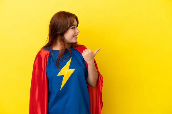 Super Hero redhead woman isolated on yellow background pointing to the side to present a product