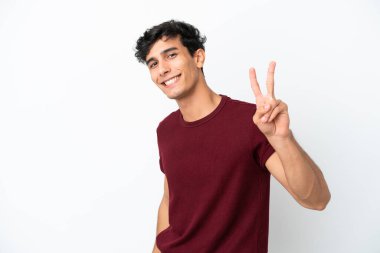 Beyaz arka planda izole edilmiş genç Arjantinli adam gülümsüyor ve zafer işareti gösteriyor.