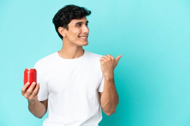 Arjantinli genç bir adam mavi arka planda izole edilmiş bir içecek tutuyor. Bir ürünü sunmak için yan tarafa bakıyor.