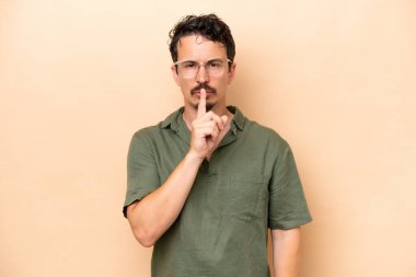 Beyazlı genç adam bej arka planda izole edilmiş. Parmağını ağzına koyduğunda sessizlik işareti gösteriyor.