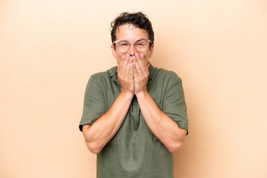 Beyazlı genç bir adam bej arka planda izole edilmiş mutlu ve gülümseyen elleriyle ağzını kapatıyor.