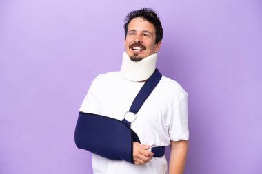 Mor arka planda izole edilmiş askı ve boyunluk takan genç beyaz bir adam gülüyor.