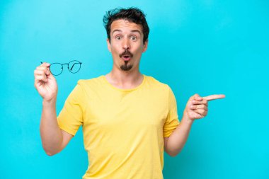 Mavi arka planda gözlüklü genç beyaz adam şaşırmış ve işaret ediyor.