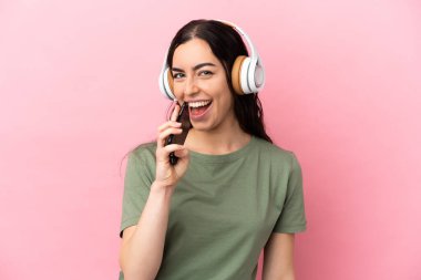 Pembe arka planda izole edilmiş genç beyaz bir kadın cep telefonuyla müzik dinliyor ve şarkı söylüyor.