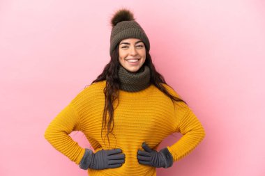 Mor arka planda izole edilmiş, kalçasında kollarıyla poz veren ve gülümseyen, kışlık şapkalı beyaz bir kız.