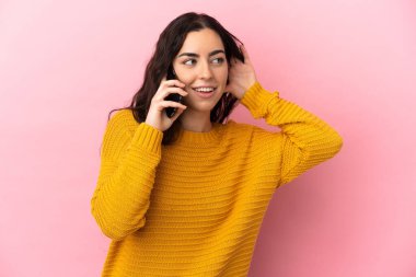 Pembe arka planda izole edilmiş cep telefonu kullanan beyaz bir kadın kulağına elini sürerek bir şeyler dinliyor.