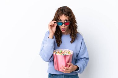 Young woman with curly hair isolated on white background surprised with 3d glasses and holding a big bucket of popcorns