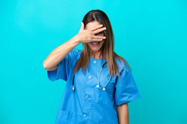 Orta yaşlı cerrah kadın mavi arka planda izole edilmiş gözleri elle kaplanmış ve gülümsüyor.