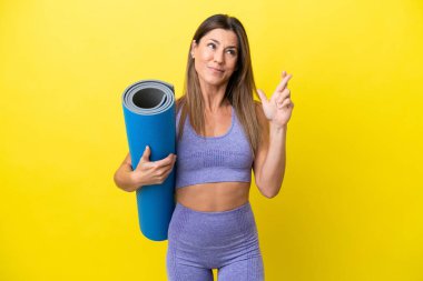 Sport woman going to yoga classes while holding a mat isolated non yellow background with fingers crossing and wishing the best