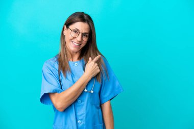 Mavi arka planda izole edilmiş orta yaşlı cerrah kadın bir ürünü sunmak için kenarı işaret ediyor.