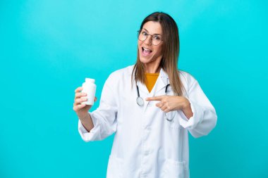 Mavi arka planda izole edilmiş orta yaşlı bir kadın doktor önlüğü giyiyor ve elinde haplar var.
