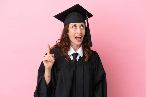 Young university graduate woman isolated on pink background pointing up and surprised
