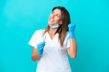 Mavi arka planda izole edilmiş orta yaşlı doktor kadın zaferi kutluyor.