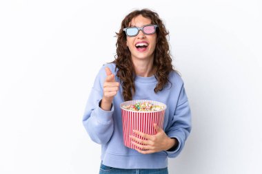 Young woman with curly hair isolated on white background with 3d glasses and holding a big bucket of popcorns while pointing front