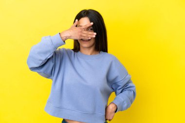 Sarı arka planda izole edilmiş genç beyaz bir kadın. Gözleri elleriyle kaplıyor ve gülümsüyor.