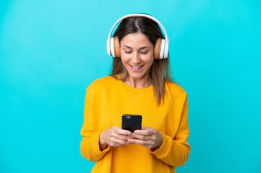 Middle age caucasian woman isolated on blue background listening music and looking to mobile