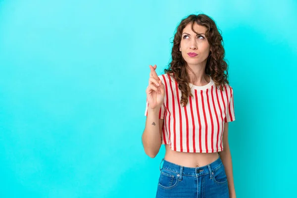 Young woman with curly hair isolated on blue background with fingers crossing and wishing the best