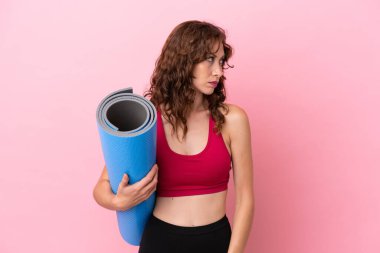 Genç sporcu kadın yoga derslerine gidiyor. Elinde pembe arka planda izole edilmiş bir minder var. Yan tarafa bakıyor.