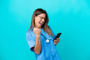 Mavi arka planda izole edilmiş orta yaşlı cerrah kadın telefon zafer pozisyonunda.