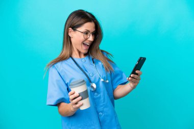 Orta yaşlı cerrah kadın mavi arka planda izole edilmiş. Elinde kahve ve cep telefonu var.