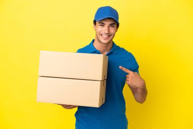 Delivery man over isolated yellow wall pointing to the side to present a product