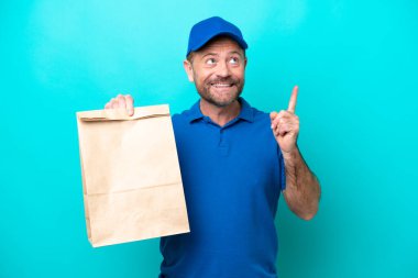 Orta yaşlı bir adam mavi arka planda izole edilmiş bir paket yiyecek alıyor. Harika bir fikre işaret ediyor.