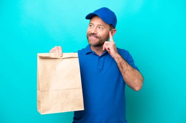 Orta yaşlı bir adam mavi arka planda izole edilmiş bir poşet yiyecek alıyor. Yukarı bakarken bir fikir düşünüyor.