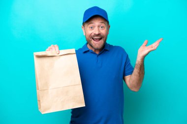 Orta yaşlı bir adam mavi arka planda izole edilmiş bir paket yiyecek alıyor ve şok olmuş bir yüz ifadesi var.