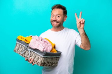 Mavi arka planda izole edilmiş çamaşır sepetini tutan orta yaşlı kadın gülümsüyor ve zafer işareti gösteriyor.