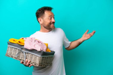 Orta yaşlı bir kadın mavi arka planda bir çamaşır sepetini tutarken sürpriz bir ifadeyle yan tarafa bakıyor.