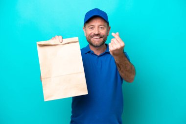 Orta yaşlı bir adam mavi arka planda izole edilmiş bir paket yiyecek alıyor ve para hareketi yapıyor.