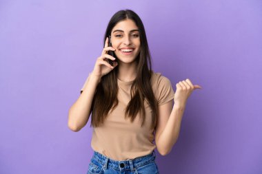 Mor arka planda izole edilmiş cep telefonu kullanan beyaz bir kadın bir ürünü sunmak için yan tarafı işaret ediyor.
