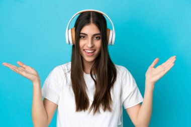 Mavi arka planda izole edilmiş genç beyaz kadın şaşırdı ve müzik dinliyor.