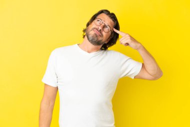 Kıdemli Hollandalı adam sarı arka planda izole edilmiş. Delilik jesti yapıyor. Parmağını kafaya koyuyor.