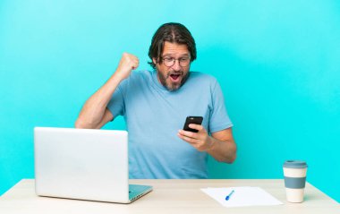Masada dizüstü bilgisayarı olan kıdemli Hollandalı adam mavi arka planda izole edilmiş, şaşırmış ve bir mesaj gönderiyor.