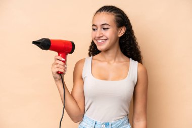 Elinde saç kurutma makinesi olan genç bir kadın bej arka planda mutlu bir ifadeyle izole edilmiş.