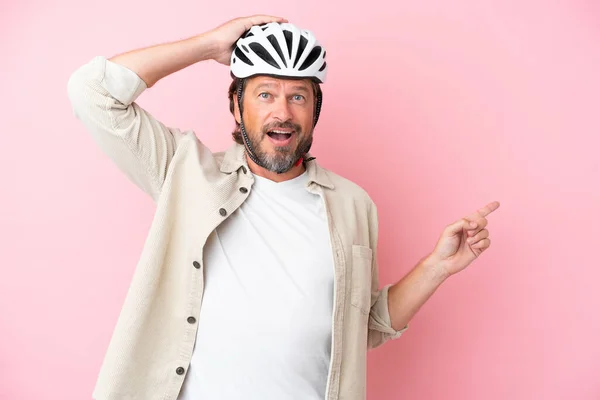 Senior dutch man with bike helmet isolated on pink background surprised and pointing finger to the side