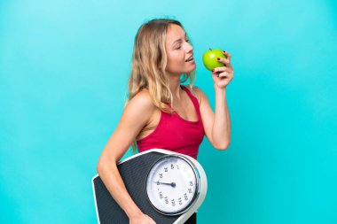 Mavi arka planda, tartı makinesi ve elmayla izole edilmiş genç bir Rus kadın.