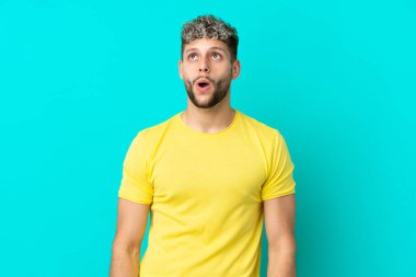 Genç yakışıklı beyaz adam mavi arka planda izole edilmiş yukarı bakıyor ve şaşırmış bir ifadeyle