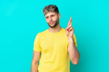 Mavi arka planda izole edilmiş genç, yakışıklı beyaz bir adam parmaklarını çapraz yapıp en iyisini diliyor.
