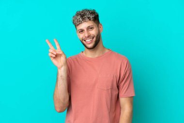 Mavi arka planda izole edilmiş yakışıklı beyaz adam gülümsüyor ve zafer işareti gösteriyor.